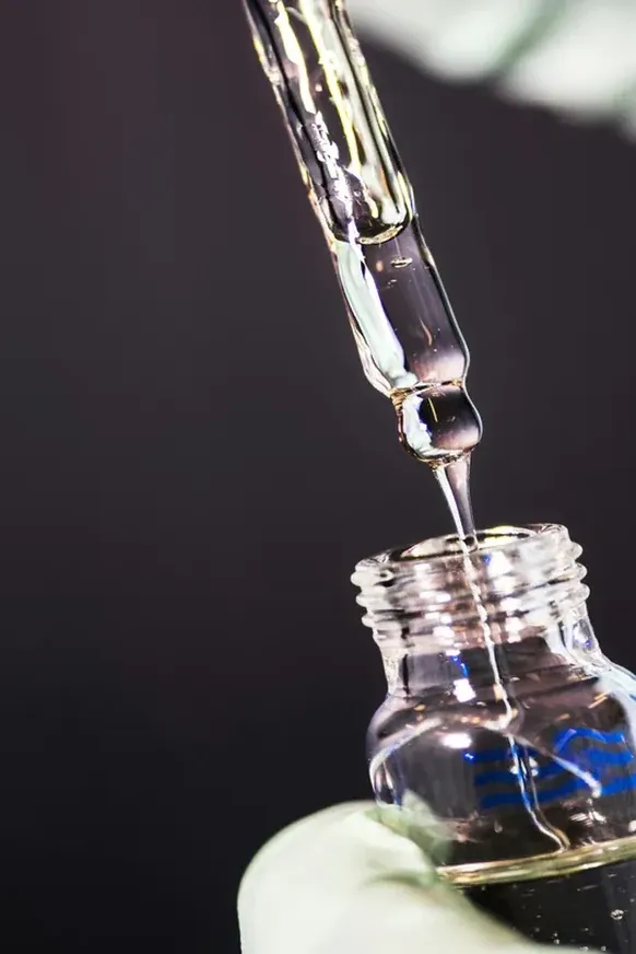 A gloved hand releases a drop from a glass pipette into a small vial against a dark background